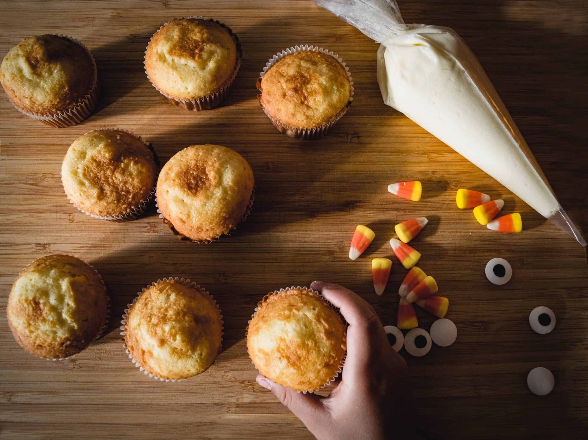 Cupcakes without frosting next to a tube of frosting and candy corn. A hand is picking up one of the cupcakes.